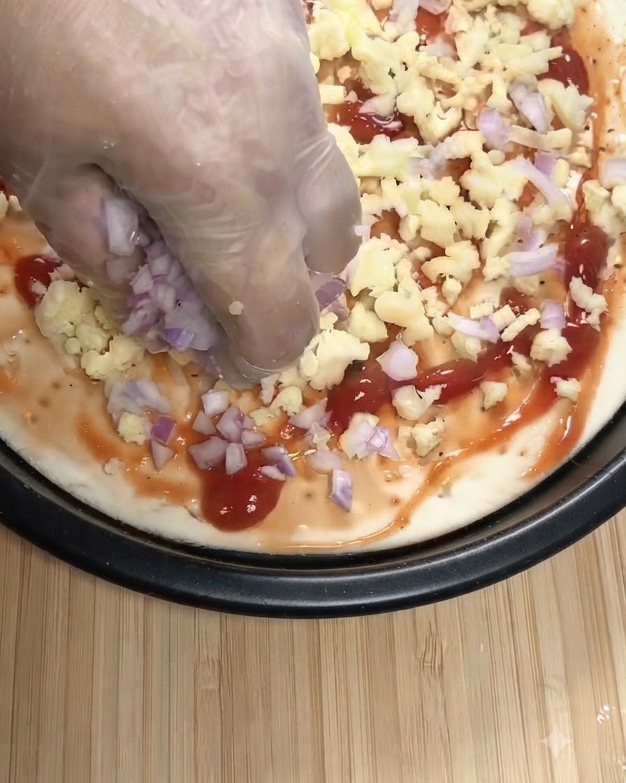 Dłoń w przezroczystej rękawiczce posypująca pokrojoną czerwoną cebulę i startą mozarellę na bazę pizzy pokrytą sosem pomidorowym.