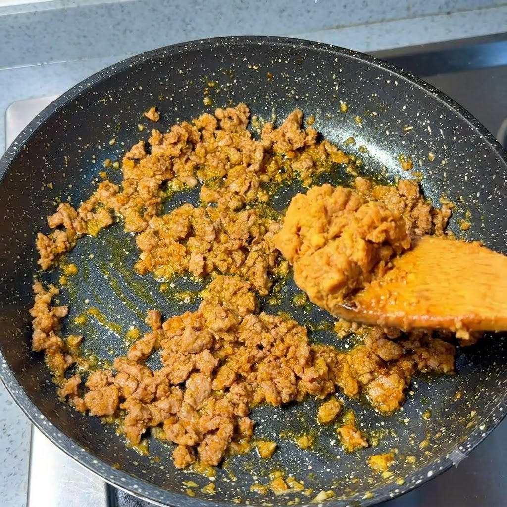 Łyżka pasty miso dodawana do usmażonej mielonej wieprzowiny na patelni.