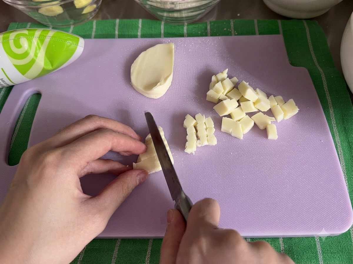 Blok sera mozzarella krojony na małe kostki nożem na desce do krojenia.