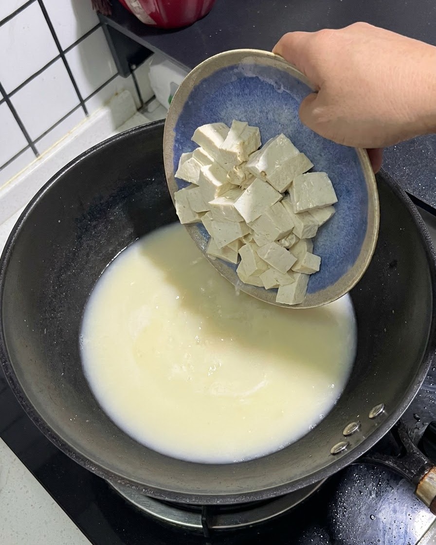 Białe kostki tofu zsuwające się z niebieskiego talerza do garnka z kremowobiałym bulionem rybnym.