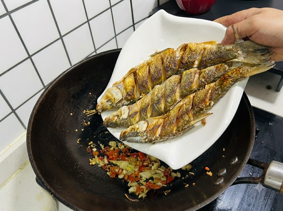 Trzy usmażone ryby zsuwane z białego talerza z powrotem do woka zawierającego podsmażony czosnek, imbir i chili.