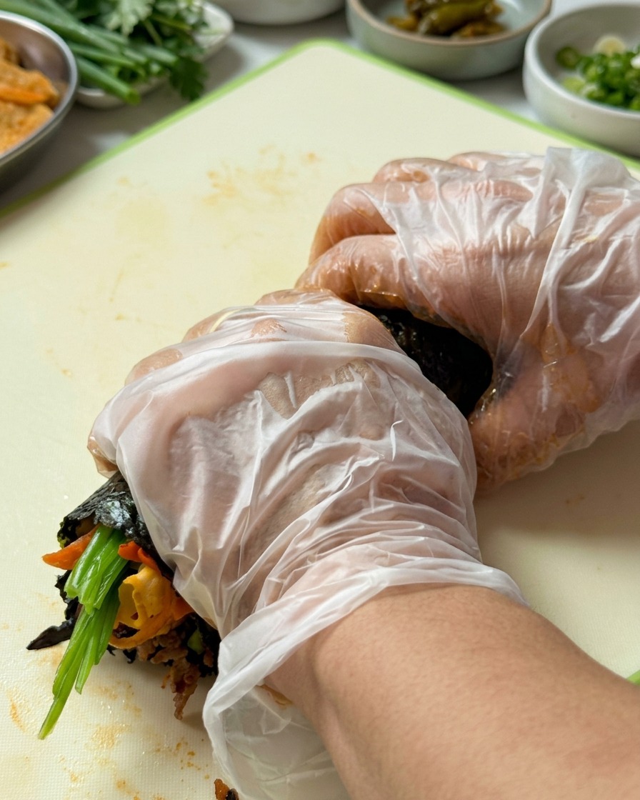 Ręce w rękawiczkach mocno zwijające gimbap od dolnej krawędzi ku górze.