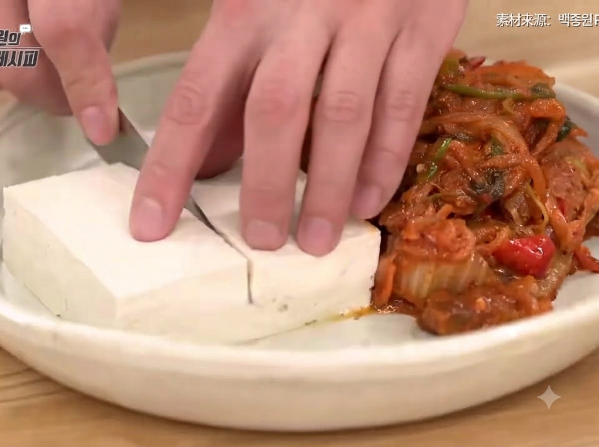 Ręka krojąca nożem zblanszowane tofu na talerzu obok porcji podsmażonego kimchi z mięsem.