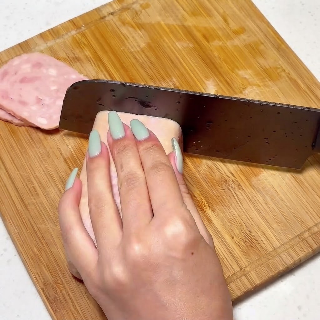 Luncheon meat krojony równomiernie na drewnianej desce za pomocą noża.