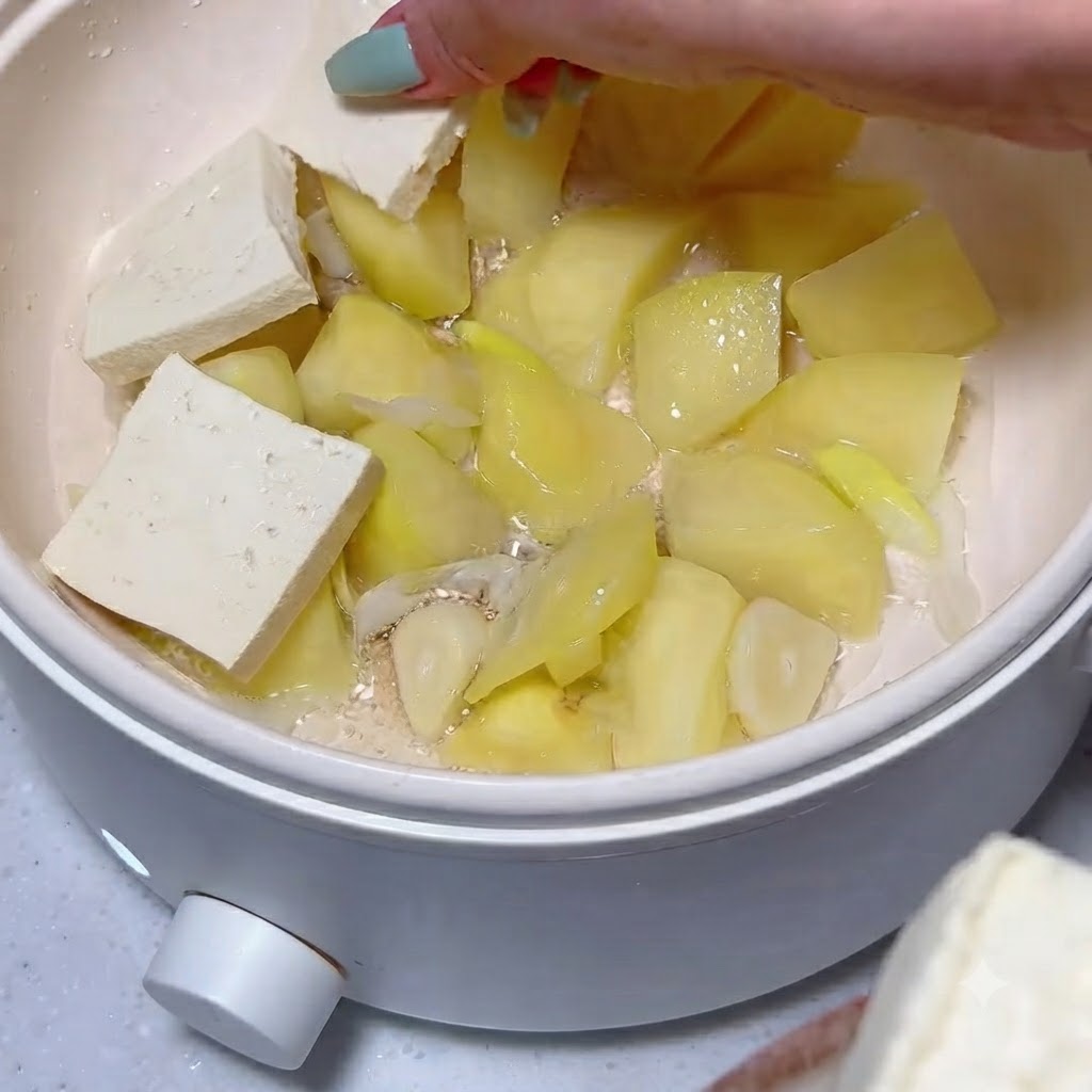 Plastry tofu ułożone schludnie na warstwie ugotowanych kawałków ziemniaków i czosnku w białym garnku.