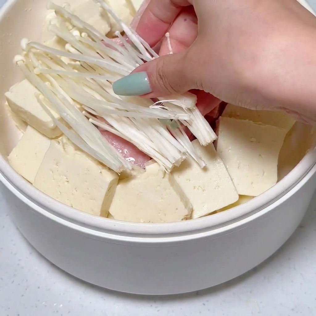 Grzyby enoki umieszczane w garnku pośród ułożonego tofu i plastrów luncheon meatu.