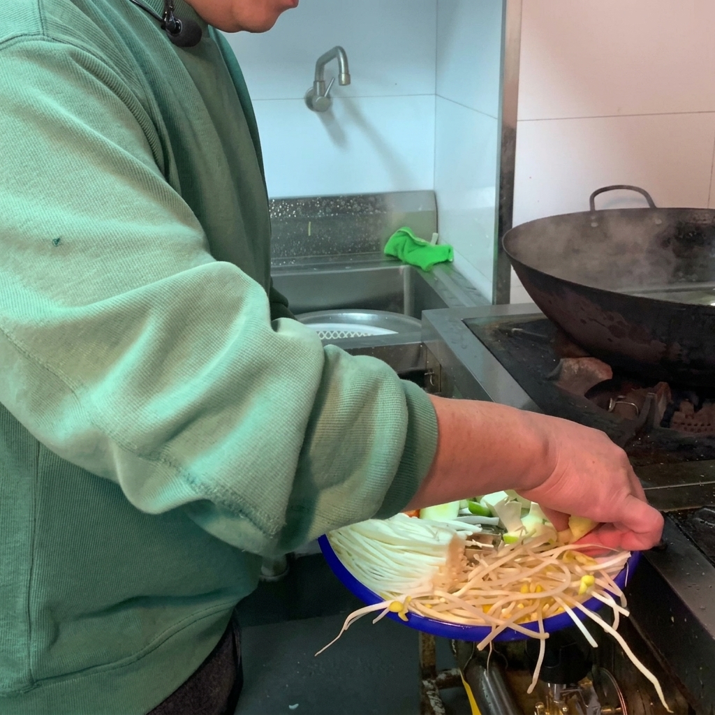 Różnorodne świeże warzywa i białe tofu wrzucane do ciemnego glinianego garnka wypełnionego wodą.