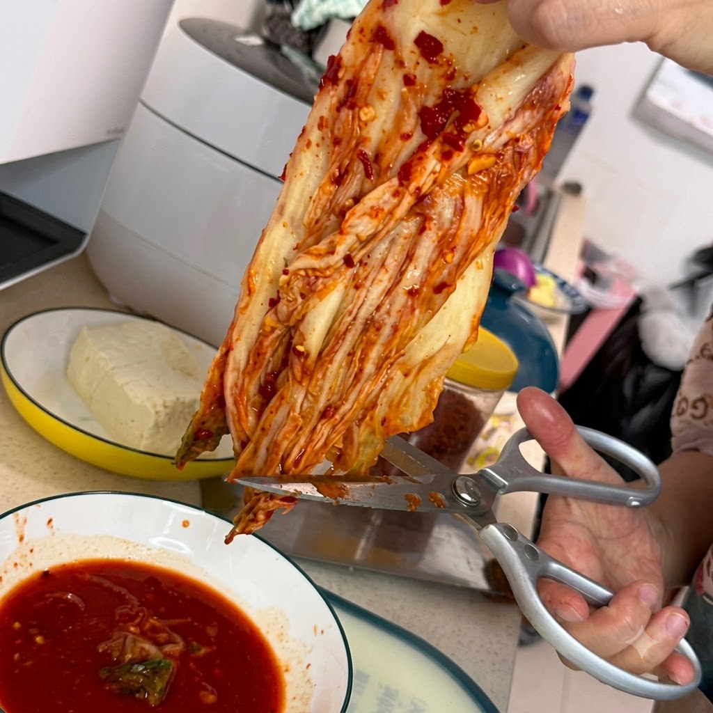 Para srebrnych nożyczek kuchennych tnących kawałek czerwonego kimchi nad białym talerzem wypełnionym pikantnym czerwonym sosem.