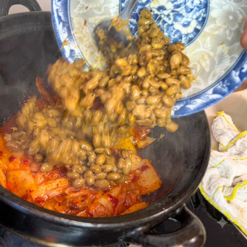 Sfermentowana soja zsuwająca się z niebiesko-białego ozdobnego talerza do ciemnego garnka wypełnionego skwierczącym kimchi.