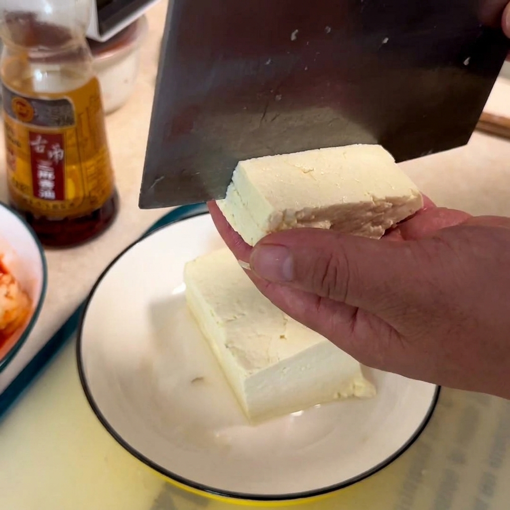 Dłoń trzymająca blok białego tofu, podczas gdy tasak kroi je w kostkę na białym talerzu.
