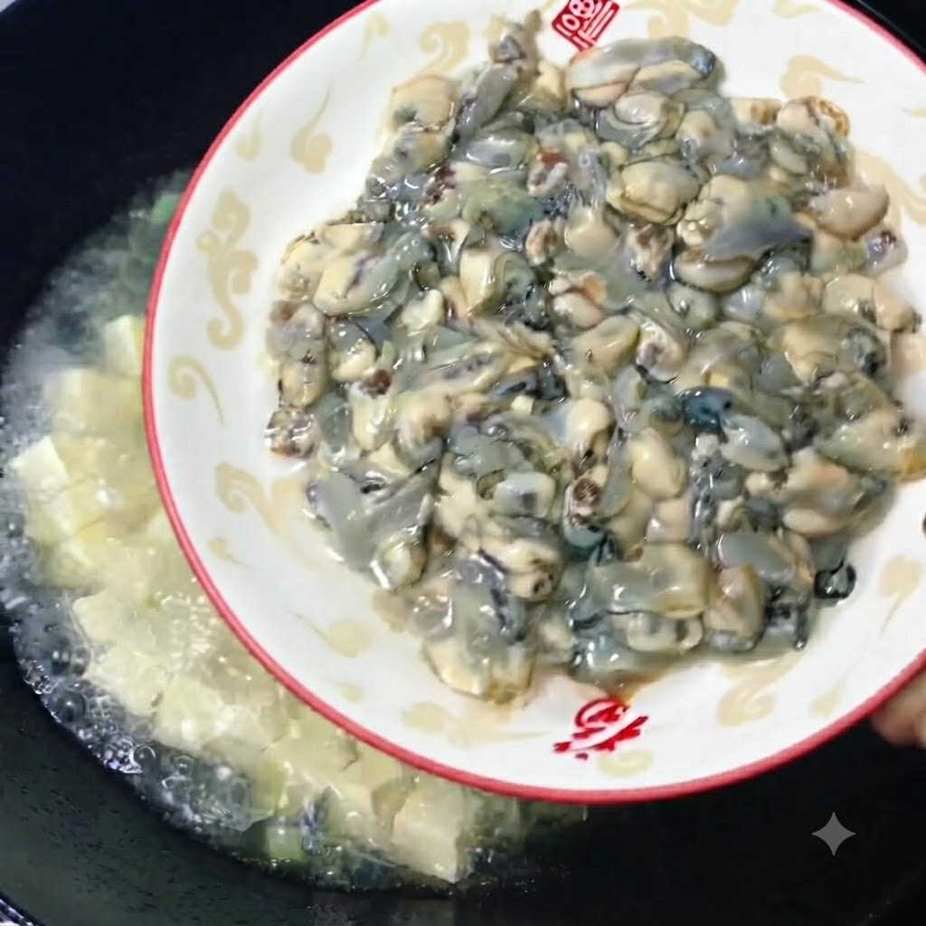 Fresh, plump raw oysters being slid from a decorative plate into a wok of simmering tofu broth.