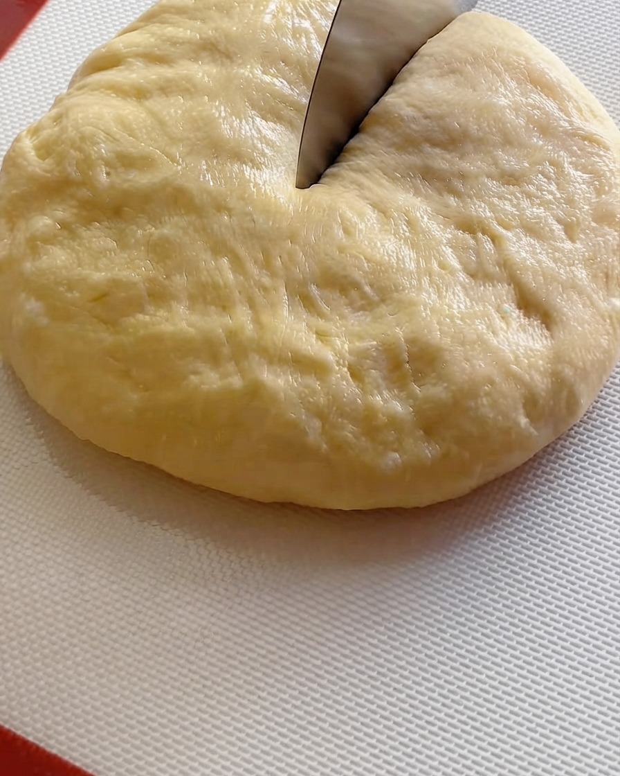 Nóż do ciasta krojący dużą kulę sfermentowanego ciasta na dziewięć porcji na białej macie.