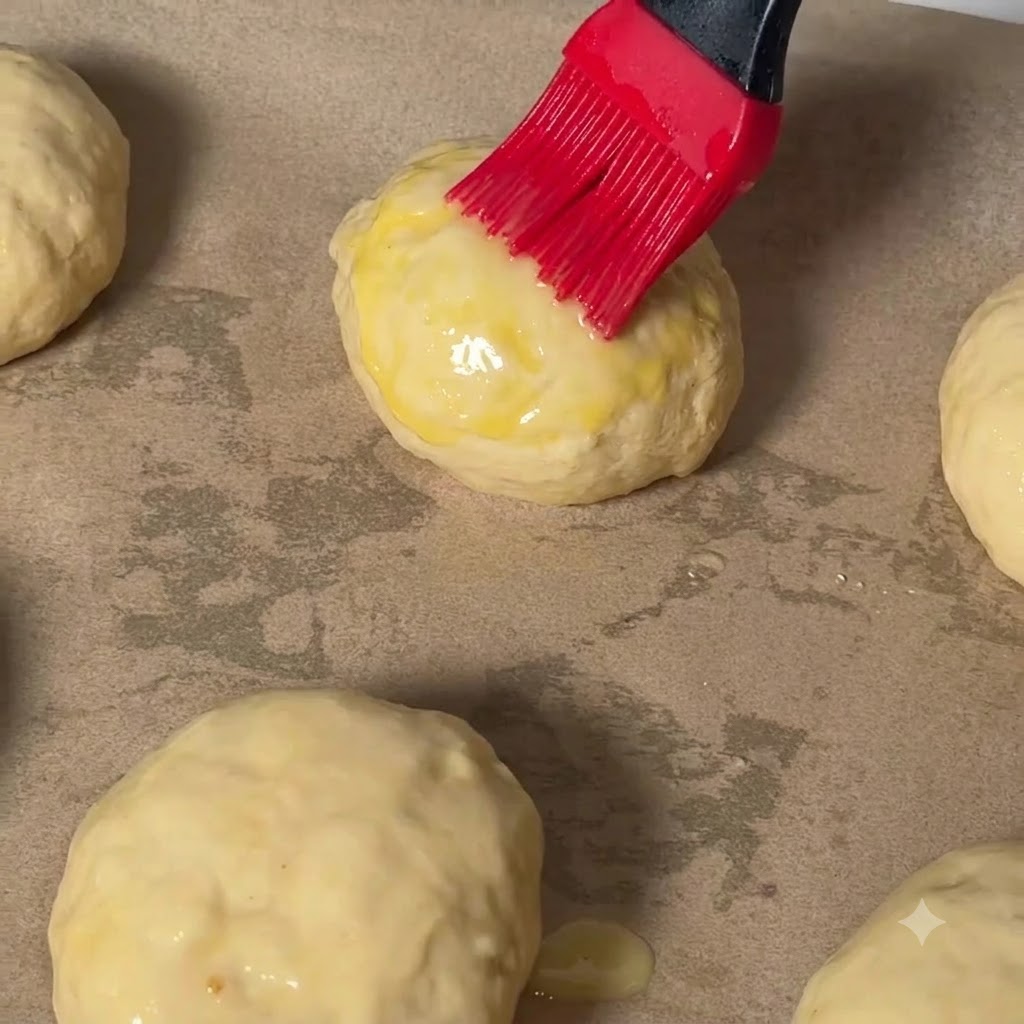 Czerwony pędzel cukierniczy nakładający wash jajeczny na wierzch kulki ciasta cheeseburgerowego na papierze do pieczenia.