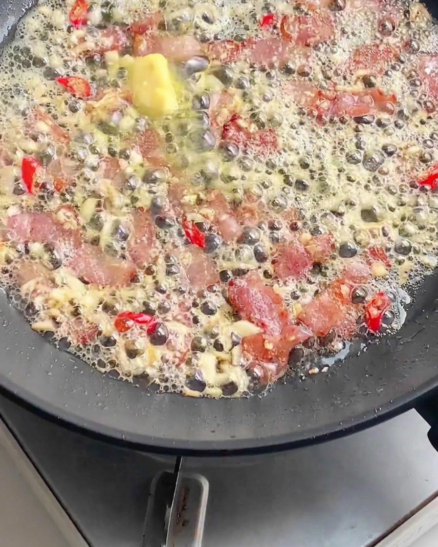Ugotowany makaron dodawany do patelni wypełnionej skwierczącym boczkiem, rozpuszczonym masłem, posiekanym czosnkiem i czerwonym chili.