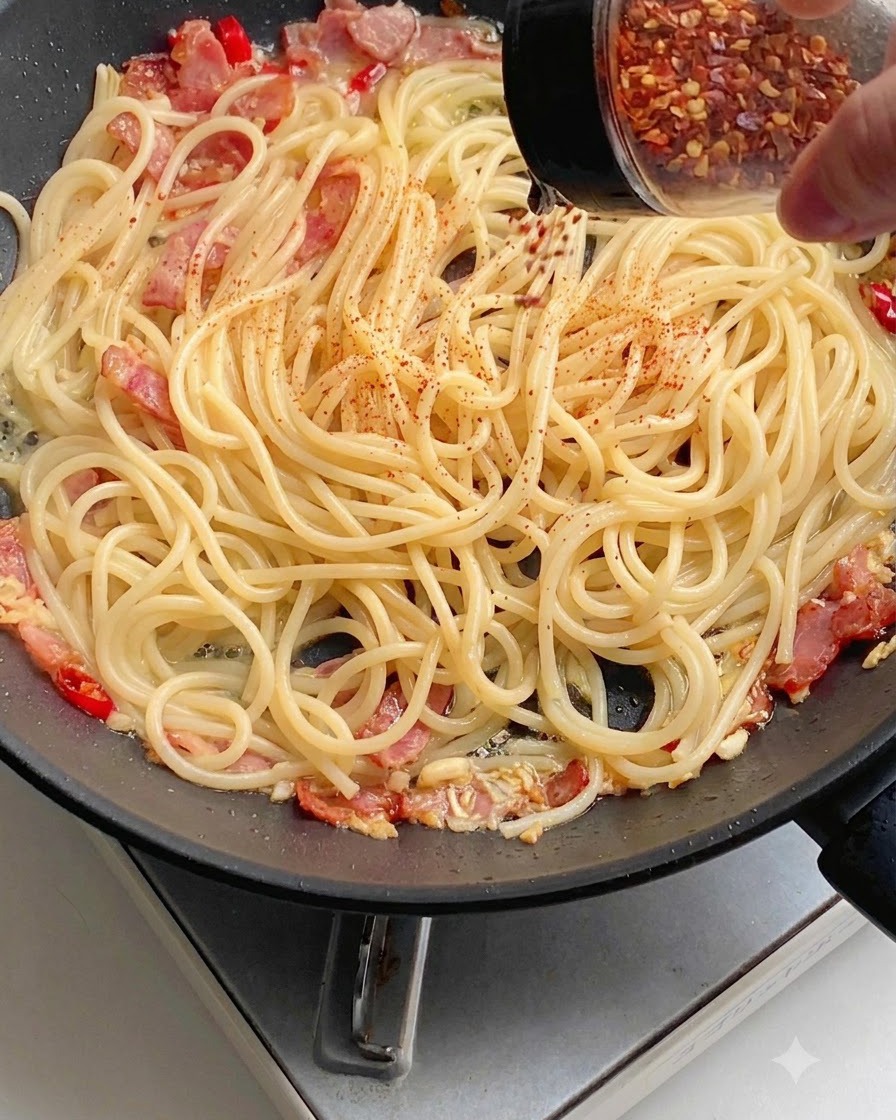 Czerwona papryka w proszku posypywana równomiernie na spaghetti na patelni.