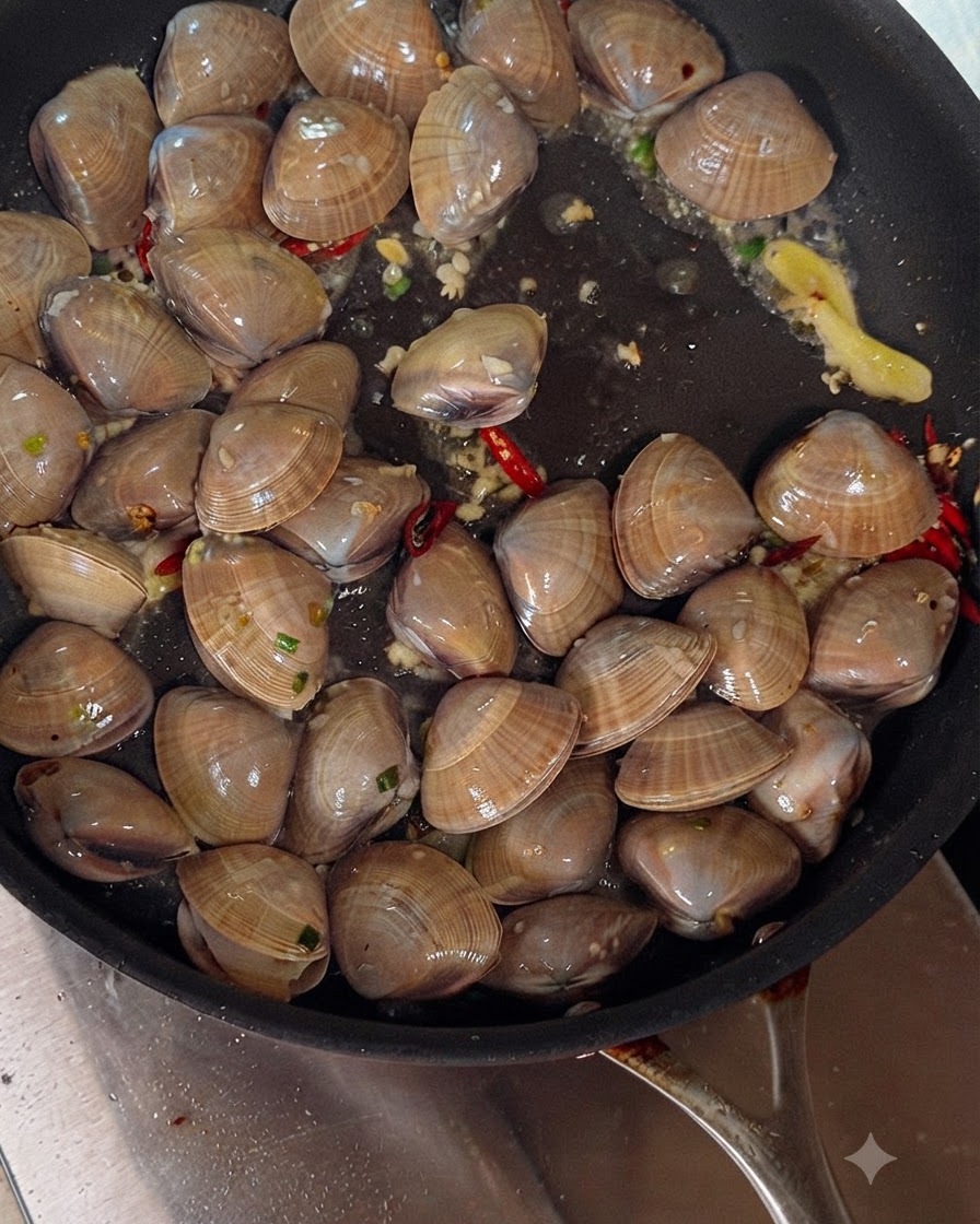 Białe wino wlewane z butelki na syczącą patelnię wypełnioną małżami, czosnkiem i papryčkami chili.