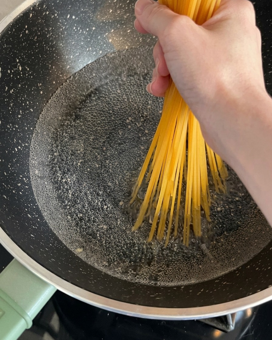 Suche nitki spaghetti trzymane przez dłoń i wkładane do bulgoczącego garnka z wodą.