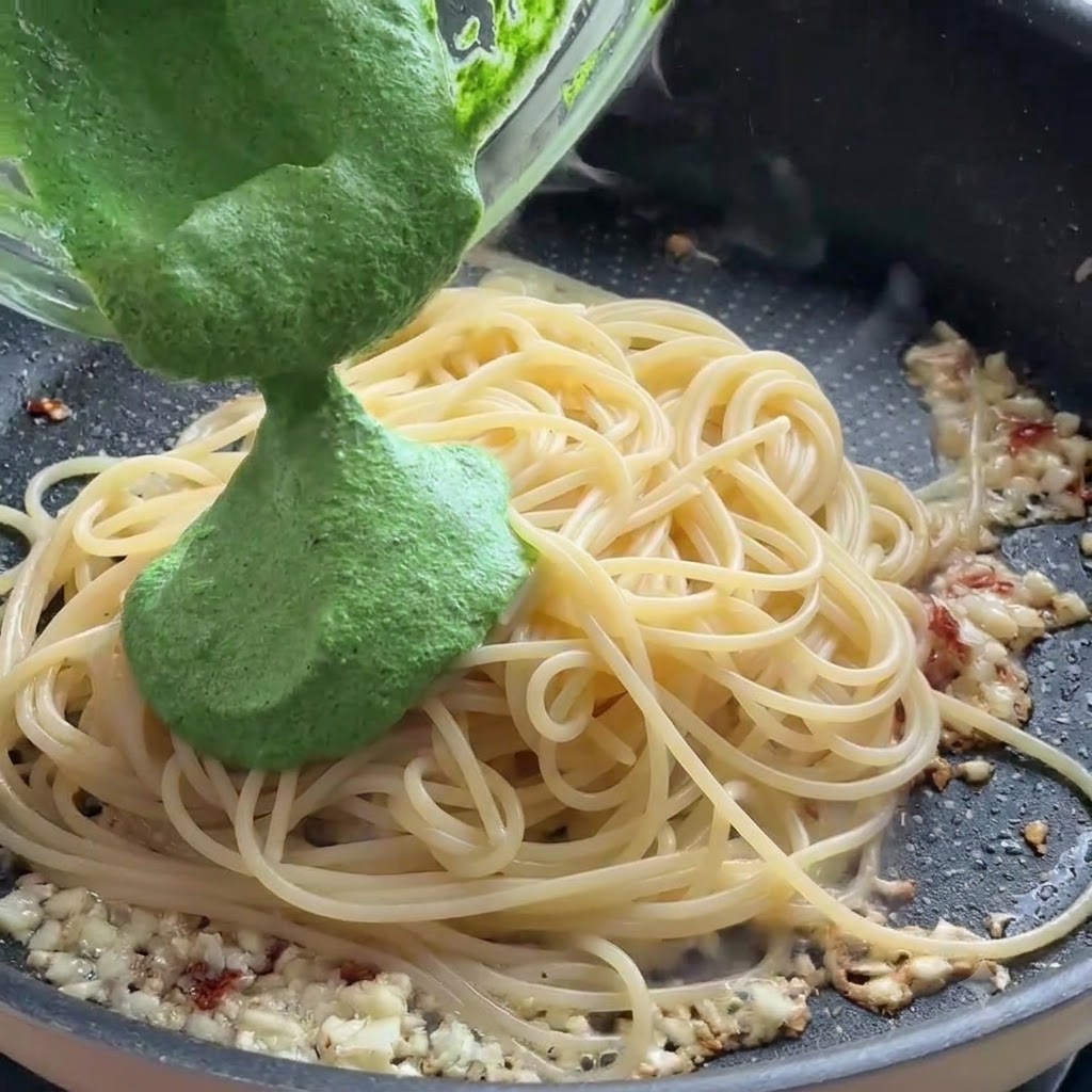 Zielone sos pesto wlewany ze szklanej miski na nitki makaronu spaghetti na patelni.