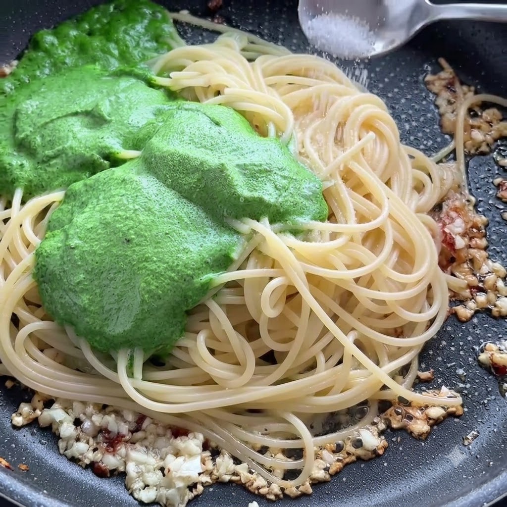 Łyżeczka soli posypywana na nitki spaghetti i zielone sos pesto na patelni.