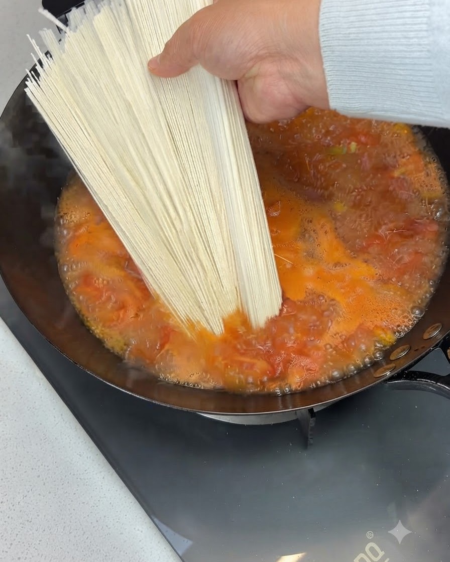 Porcja suchego makaronu trzymana nad bulgoczącą zupą pomidorową w woku i do niej dodawana.