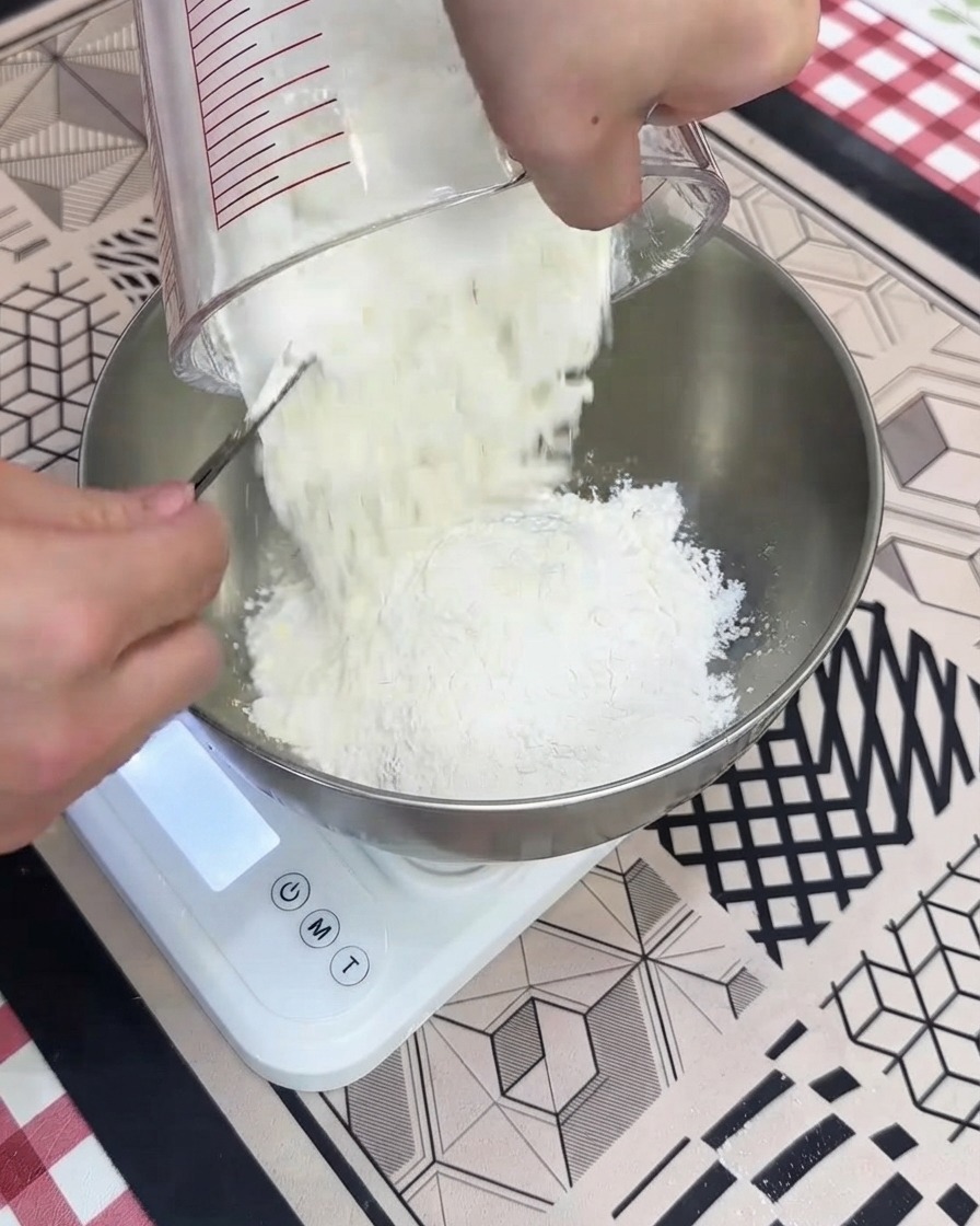Wysypywanie suchej mąki z miarki do metalowej miski na białej wadze kuchennej.