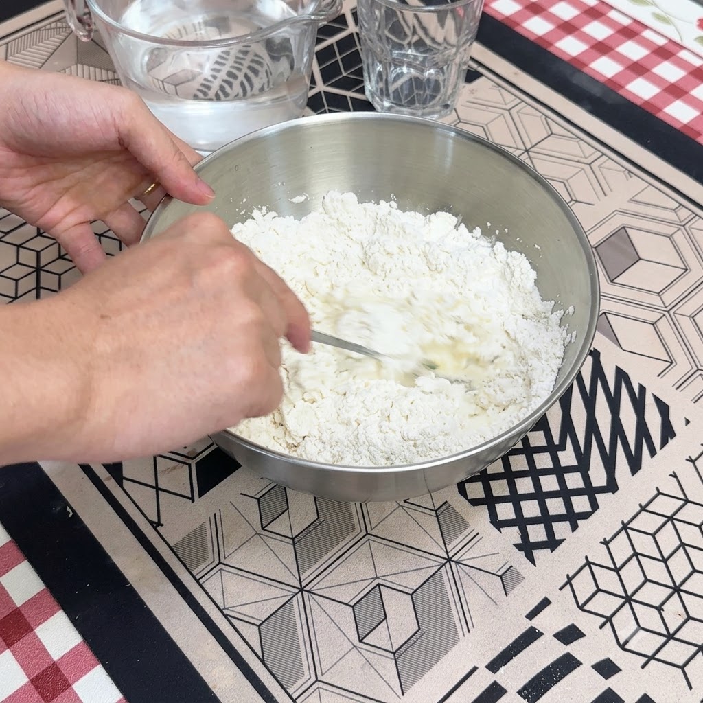 Ręka mieszająca łyżką wodę z suchą mąką w metalowej misce.