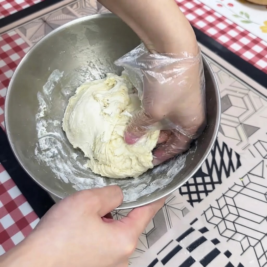 Dłoń w rękawiczce wyrabiająca wilgotne ciasto na pizzę w misce ze stali nierdzewnej.