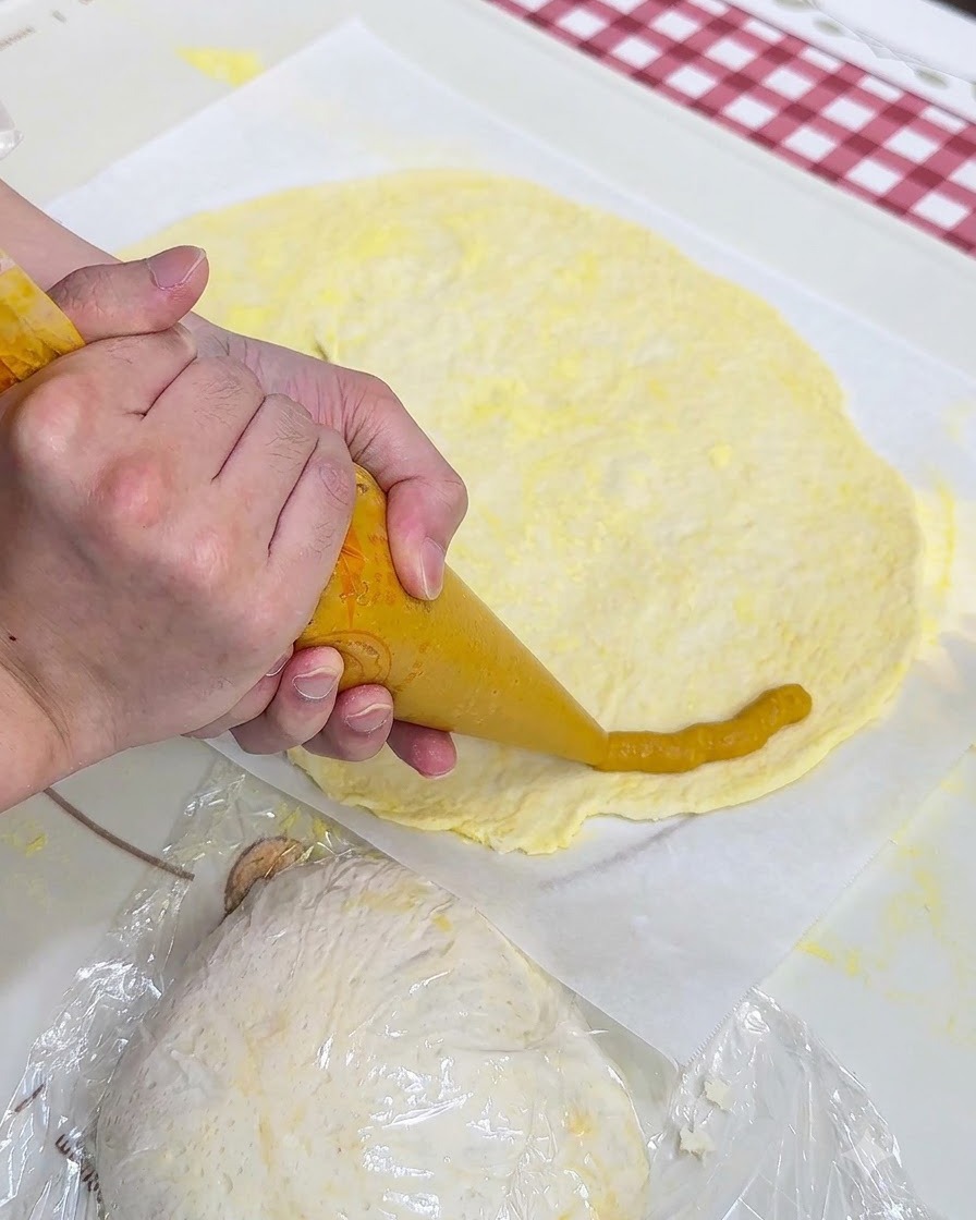 Dłonie wyciskające żółte purée z batatów z rękawa cukierniczego wokół brzegu spodu do pizzy na papierze do pieczenia.