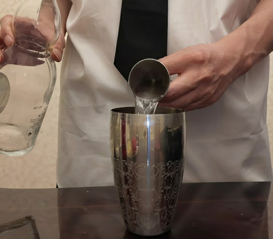 Przezroczysta tequila wlewana ze szklanej butelki do metalowej miarki trzymanej nad srebrnym szejkerem.
