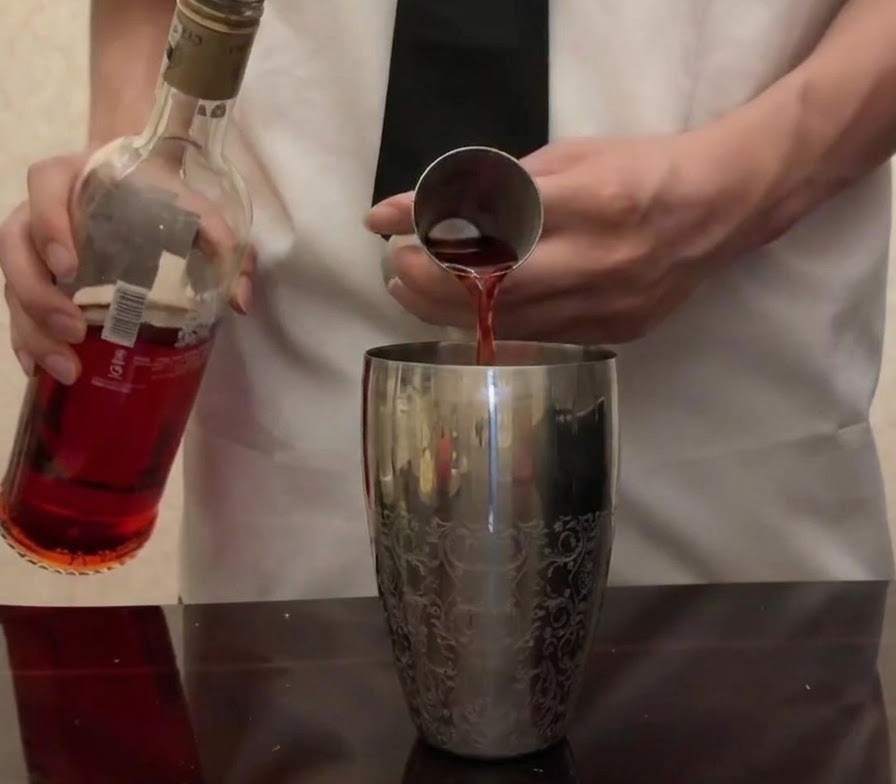 Jaskrawoczerwone Campari wlewane z butelki do metalowej miarki nad srebrnym szejkerem.