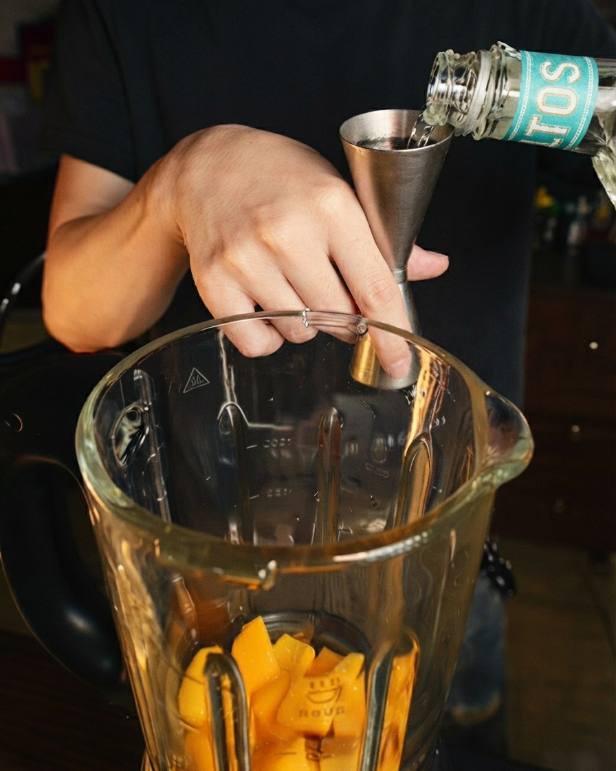 Przeźroczysty alkohol wlewany ze stali nierdzewnej miarki do dzbana blendera z kawałkami świeżego mango.