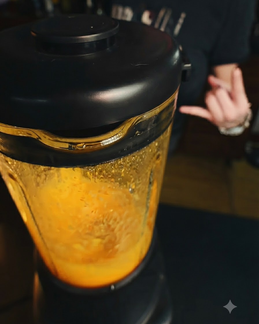 Działający blender z żywą pomarańczową masą mango w środku oraz dłonią wykonującą gest w tle.