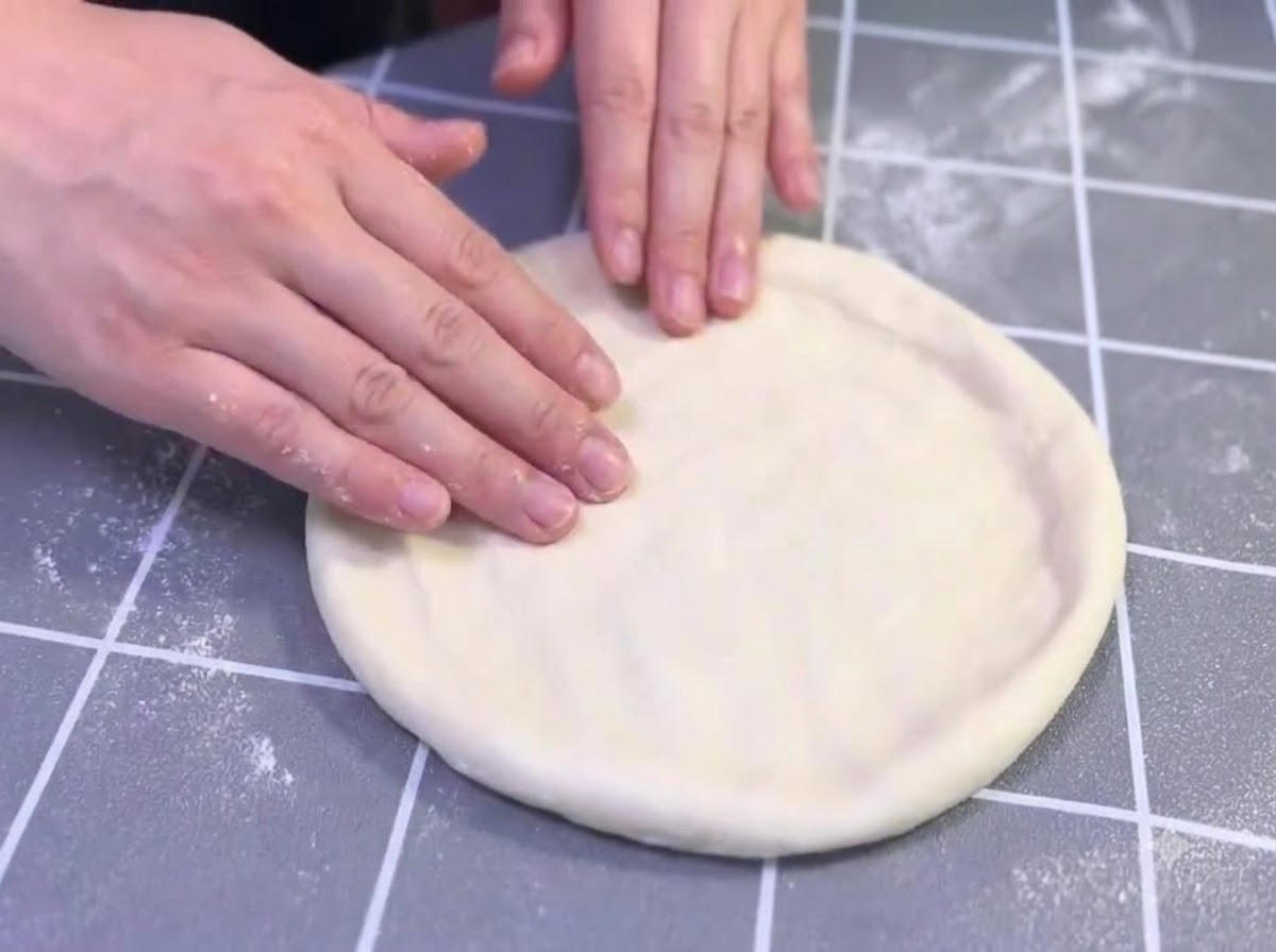 Dłonie delikatnie rozciągające i formujące naoliwiony krążek ciasta do pizzy na szarej powierzchni w kratkę.