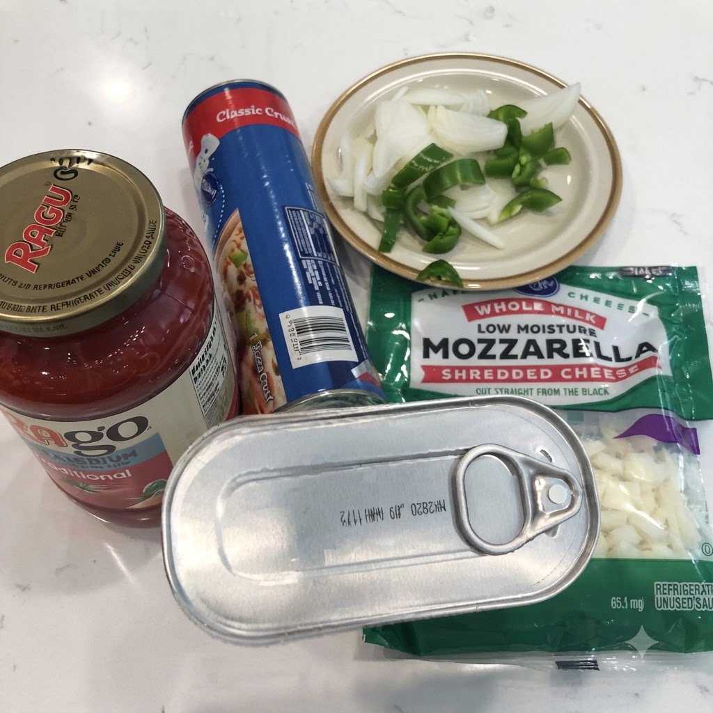 Puszka anchois, słoik sosu marinara, puszka ciasta do pizzy, starta mozzarella oraz pokrojona cebula i papryka na białym blacie.