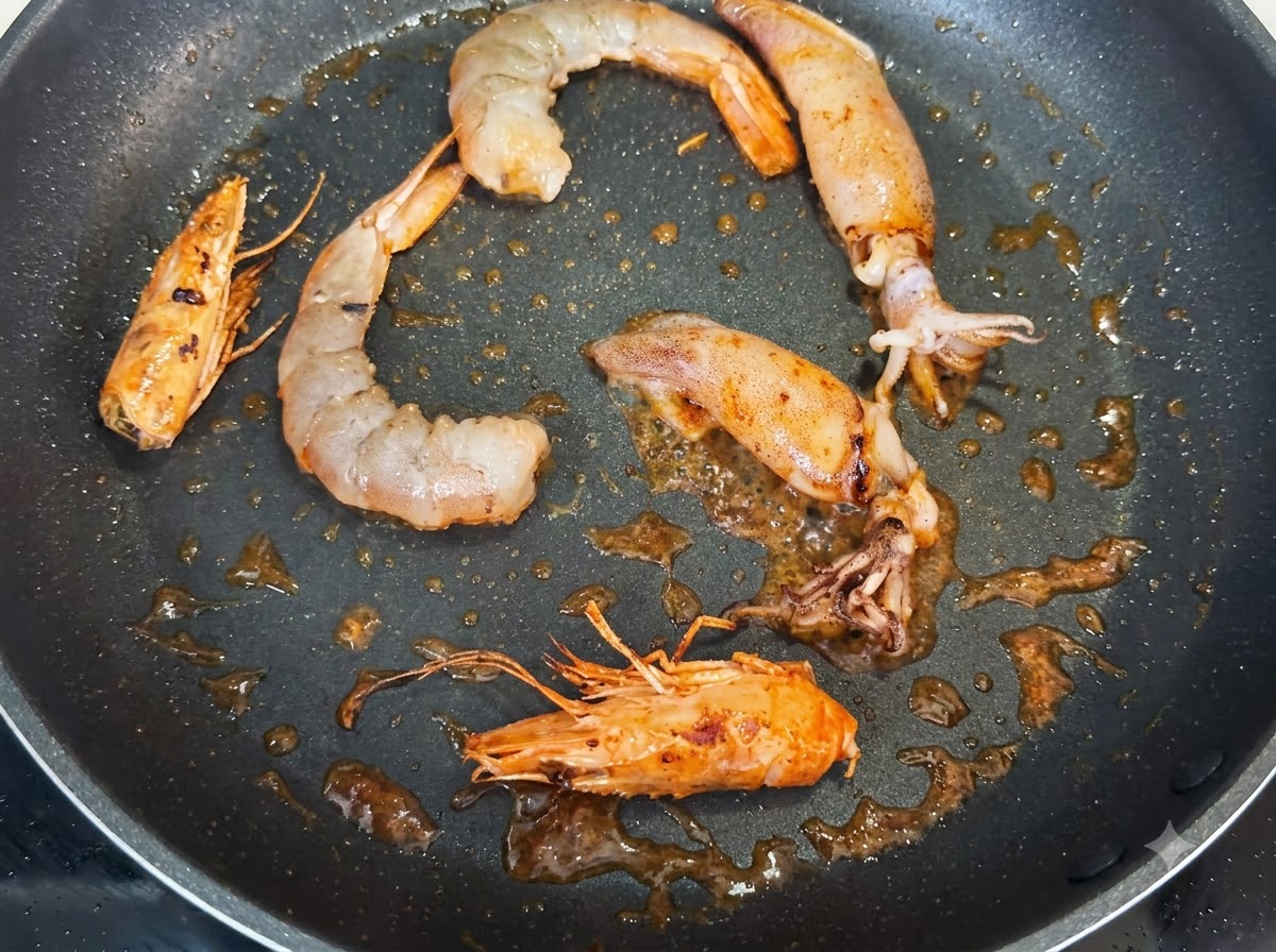 Krewetki, całe główki krewetek i kawałki kalmarów smażące się na patelni, przyprawione ciemnymi drobinkami czarnego pieprzu.