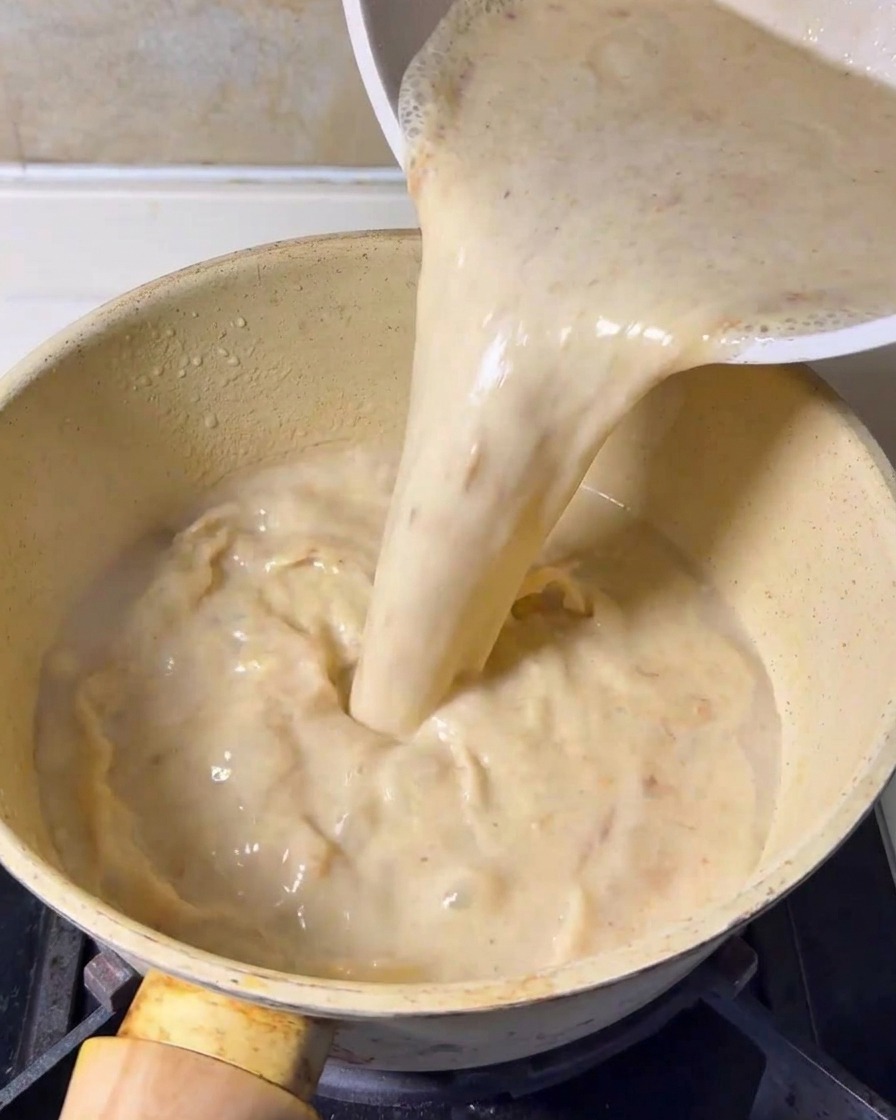 Zmiksowana kremowa baza zupy wlewana z pojemnika do żółtego garnka kuchennego na kuchence.