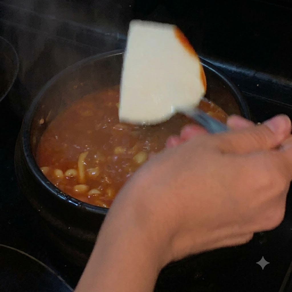 Kawałek miękkiego tofu opuszczany za pomocą łopatki do garnka z czerwoną, gotującą się zupą.