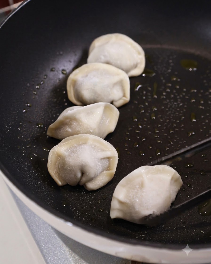 Szczypce kuchenne umieszczające surowe mrożone pierogi na ciemnej, naoliwionej płaskiej patelni obok kilku innych pierogów.
