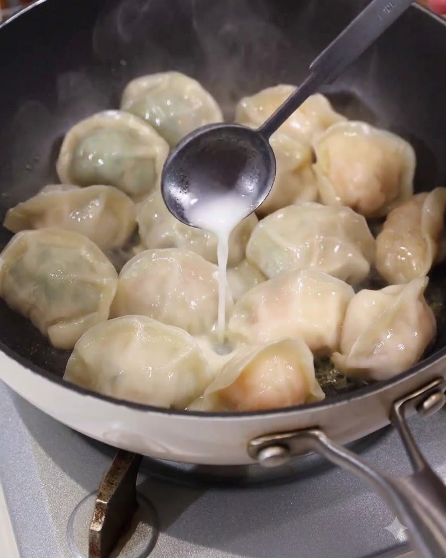 Łyżka wlewająca płynną białą zawiesinę równomiernie w przestrzenie między pierogami na gorącej patelni.