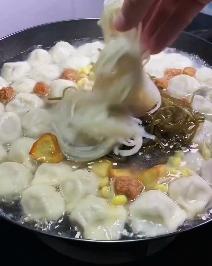 Dłoń umieszczająca posiekane zielone glony kelp i półprzezroczysty makaron sojowy w garnku z gotującymi się pierożkami i bulionem.
