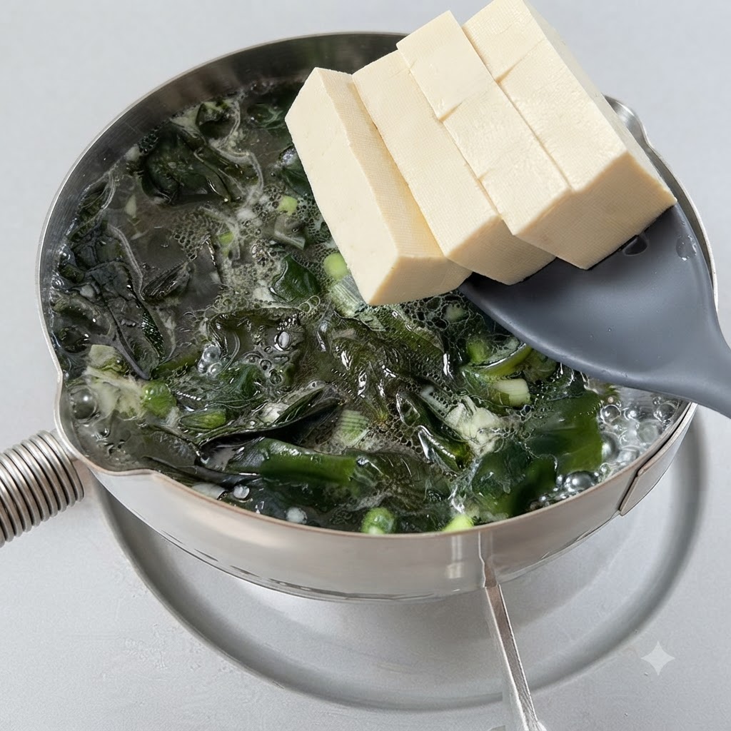 Kostki tofu ostrożnie wkładane do gotującej się zupy z glonami przy pomocy łyżki cedzakowej.