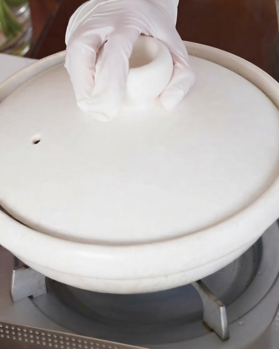 Ręka umieszczająca pokrywkę na białym ceramicznym garnku do duszenia stojącym na kuchence.