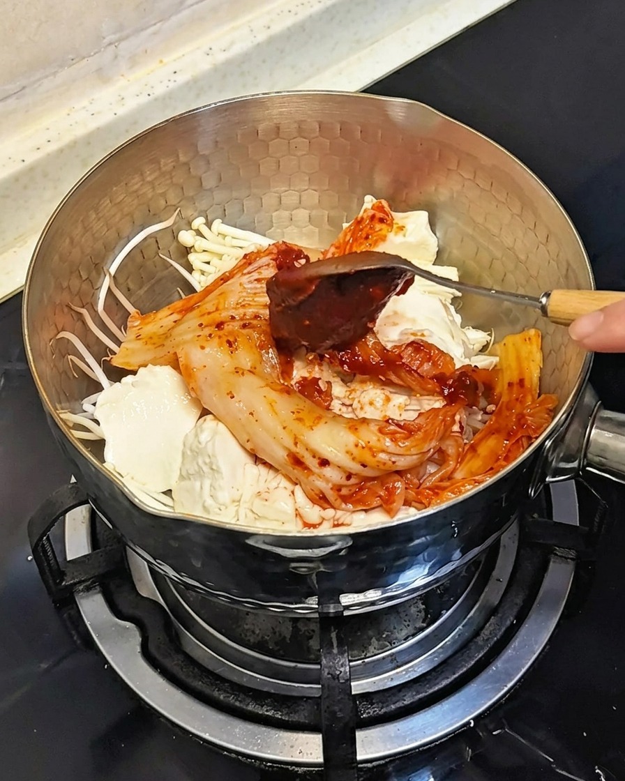 Łyżka dodająca porcję gęstej, czerwonej koreańskiej pasty chili na składniki w garnku.