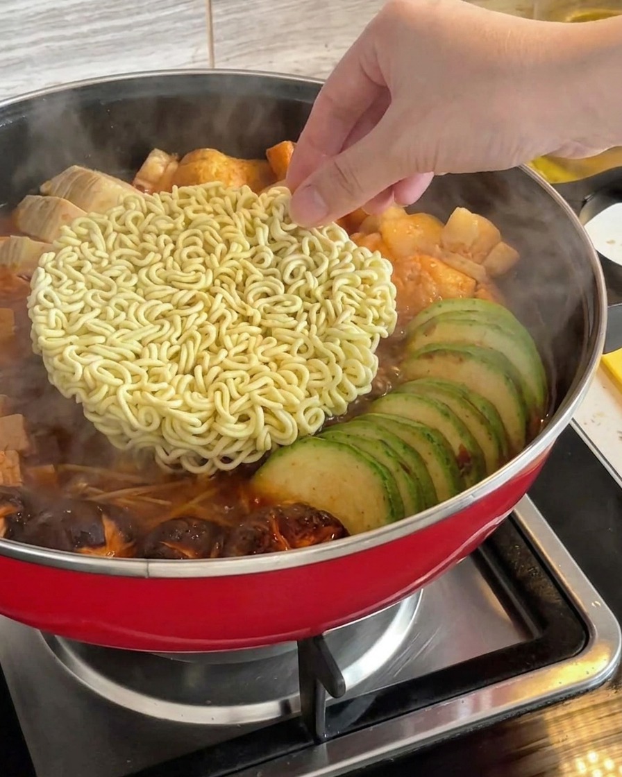 Dłoń wkładająca blok suchego błyskawicznego makaronu ramen na środek gotującego się koreańskiego gulaszu wojskowego.