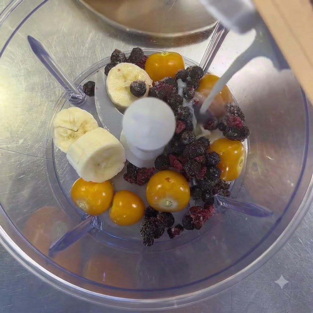 Mleko z kartonu wlewane do blendera wypełnionego miechunką, plastrami banana i owocami leśnymi.