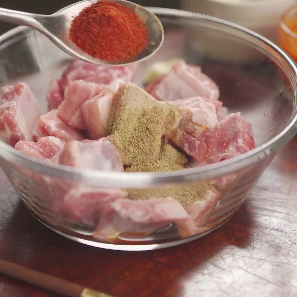 Czerwone chili w proszku na łyżce trzymanej nad miską surowych żeberek pokrytych mieszanką przypraw i ziół.