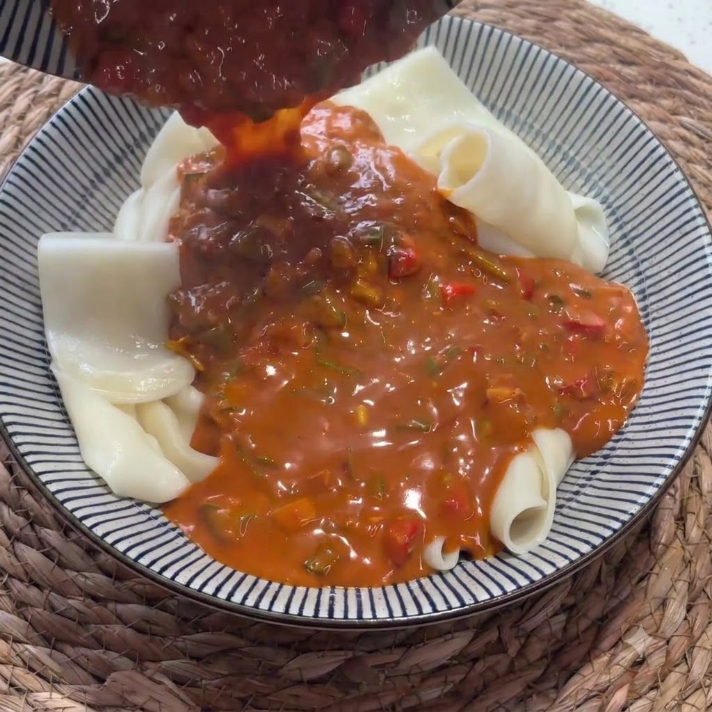 Bogaty czerwony sos wlewany chochlą na miskę wypełnioną uformowanym szerokim makaronem.