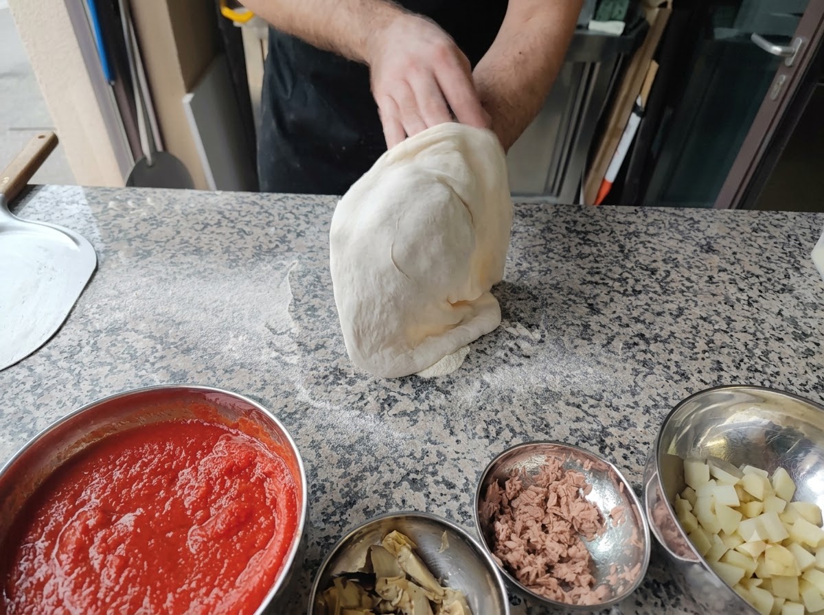 Osoba rozciągająca ciasto na pizzę w powietrzu za pomocą dłoni zwiniętych w pięści.