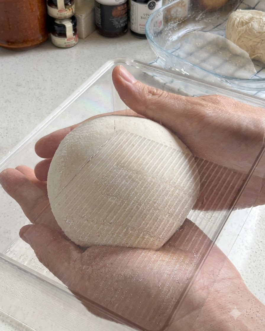 Dwie dłonie trzymające idealnie okrągłą kulę ciasta na pizzę podczas wkładania do przezroczystego plastikowego pojemnika fermentacyjnego.
