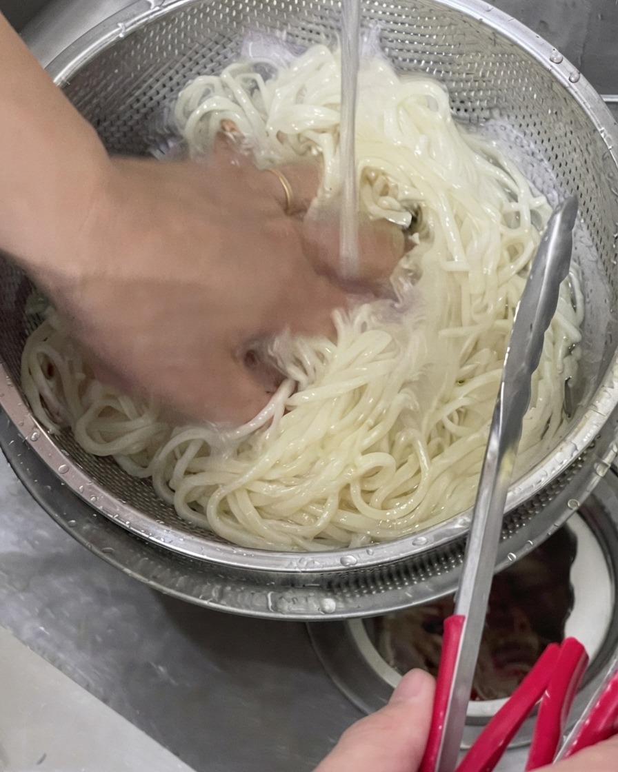Dłoń płucząca ugotowany makaron udon w metalowym sitku pod zimną bieżącą wodą.